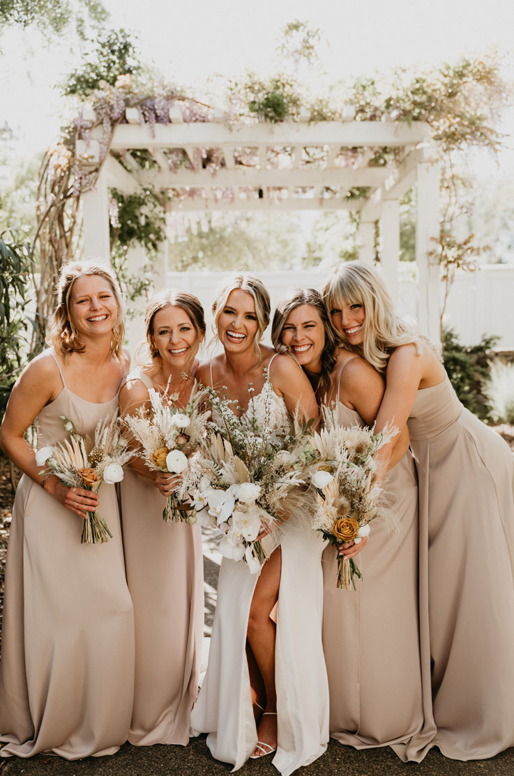 A bride in a skirt slit wedding dress and four bridesmaids smile and stand together outdoors with a pergola behind them, while holding bouquets.