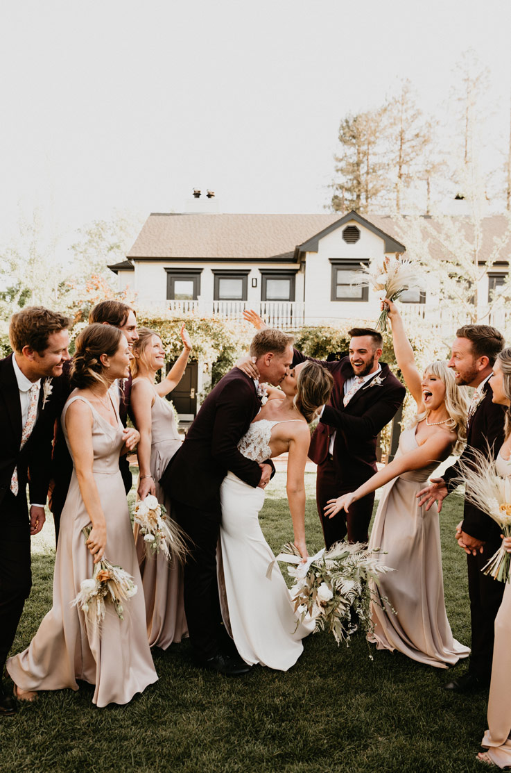 A bride in a spaghetti strap wedding dress, holding a bouquet, and a groom in a suit, kiss outdoors on a yard in front of a house, while their wedding party cheers around them.