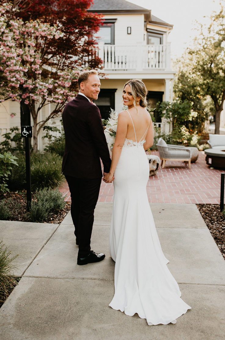 A bride in a fitted wedding dress and a groom in a suit stand on a path together, in front of a house and patio, while looking over their shoulders and smiling.