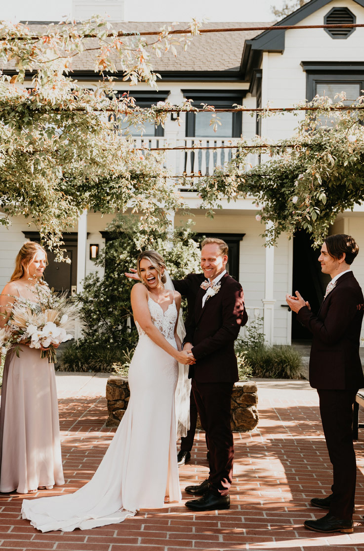 A bride in a V-neck wedding dress and a groom in a suit hold hands and laugh while standing on a patio outdoors, in front of a house, with a bridesmaid and a groomsman.