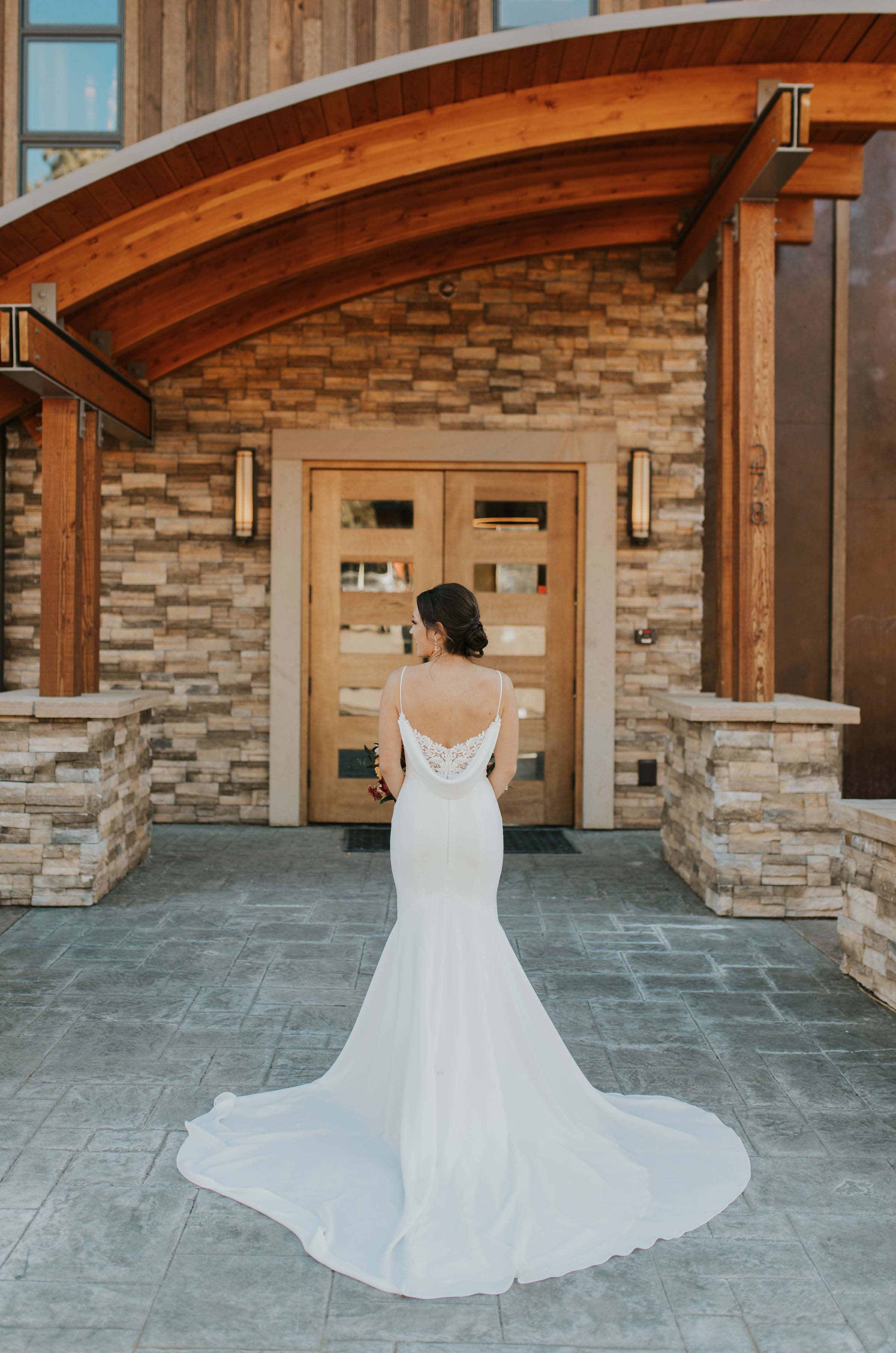 Bride in a low back crepe wedding dress with lace detail, thin straps and a fit and flare skirt, poses by venue doors.