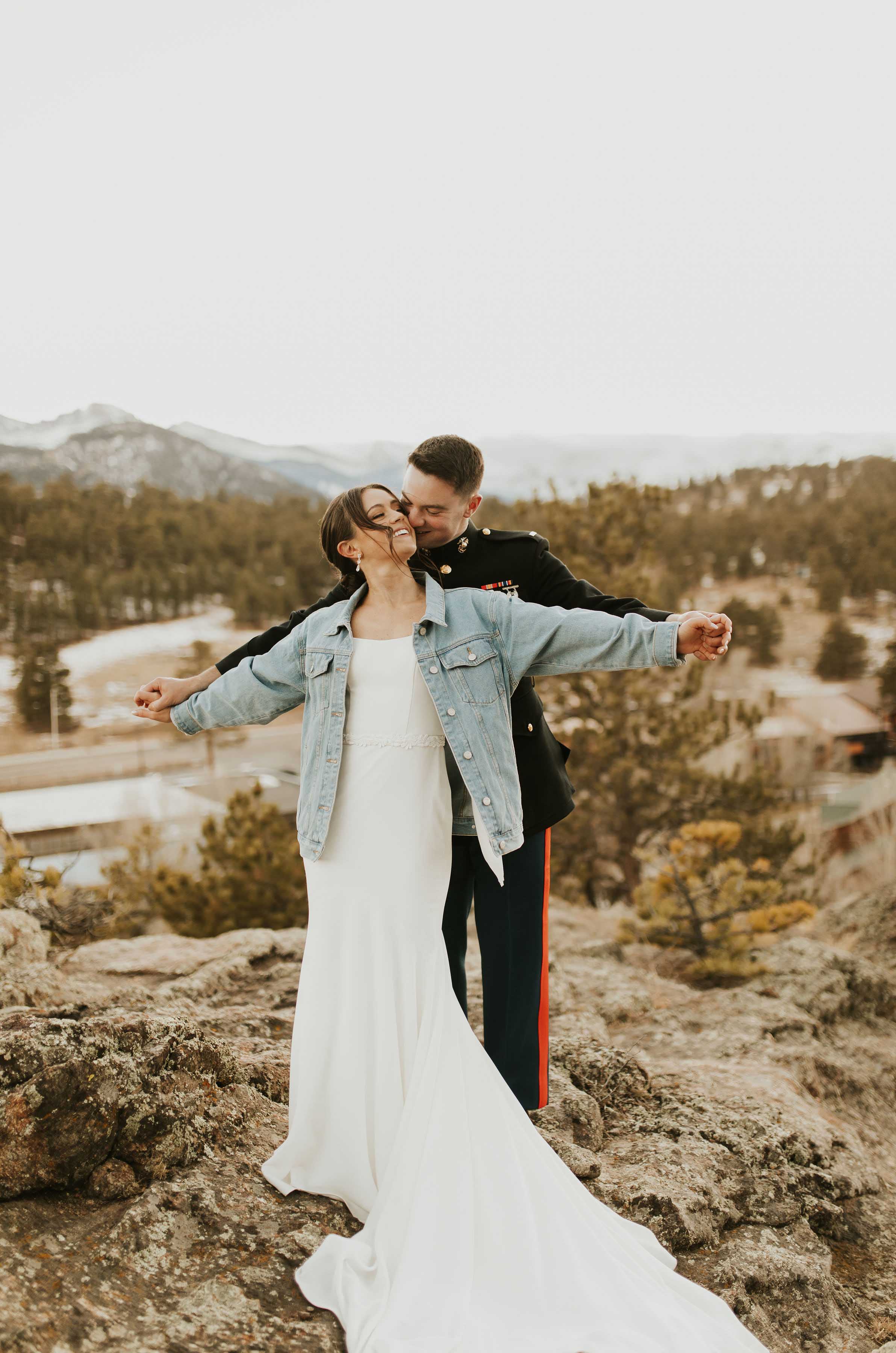 Bride in a crepe scoop neck gown with fit and flare skirt and a denim jacket, poses with groom on a mountain overlook.