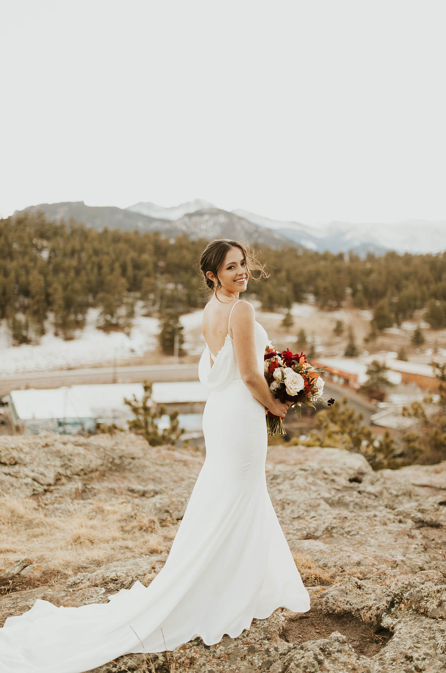 Bride looks over her shoulder, in a crepe fit and flare gown with a cowl back and thin straps, holds a bouquet on a mountain edge.