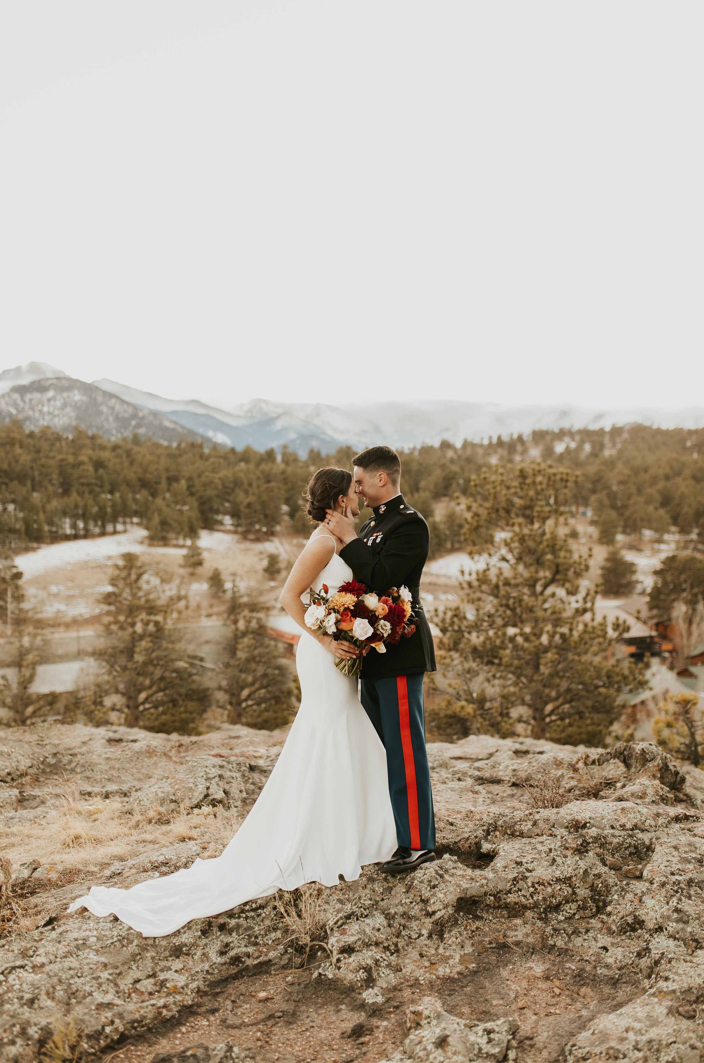 Bride in a crepe fit and flare gown with spaghetti straps and a scoop neck kisses groom in uniform on mountain ridge. 