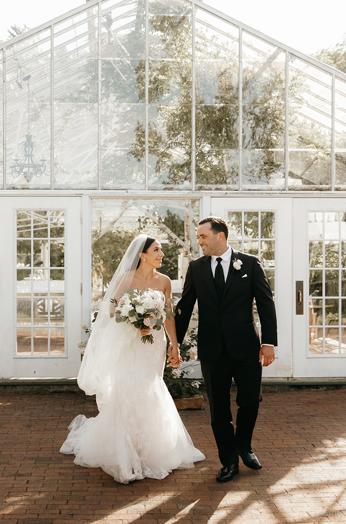 Bride in a strapless tulle gown with sequin lace and a veil, walks hand in hand with groom, holding a bouquet at greenhouse doors.