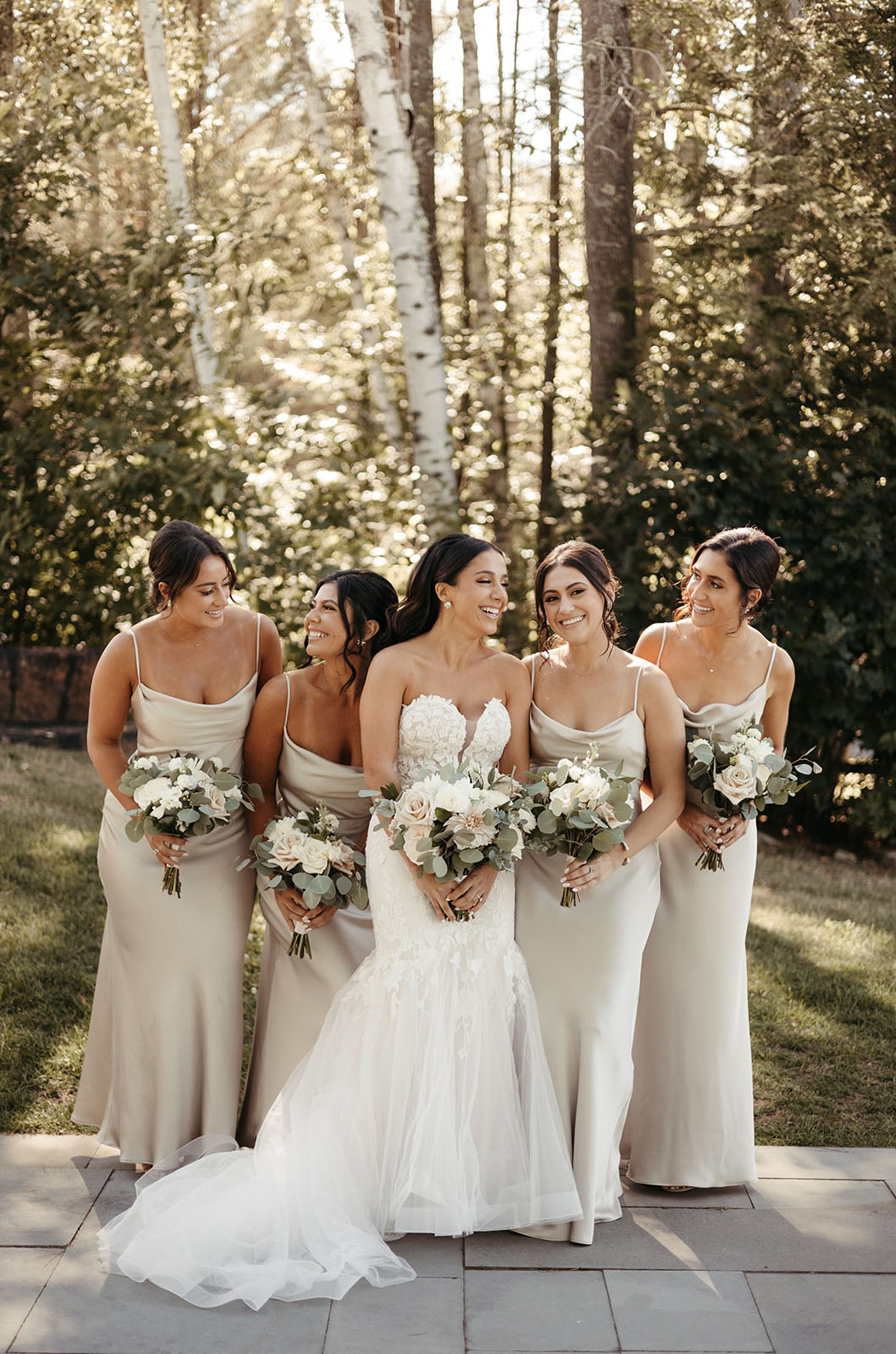 Bride in a sweetheart neck lace and sequin tulle gown smiles next to bridesmaids in satin dresses outdoors, holding bouquets.