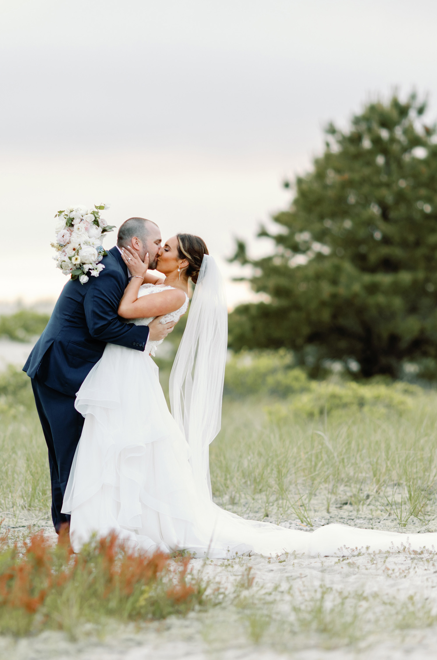 Groom in a navy suit kisses bride in a sleeveless lace bodice wedding dress with a layered tulle skirt and a veil, outdoors.
