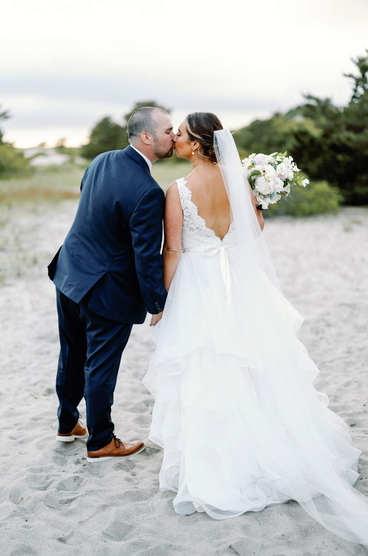 Groom kisses bride in a lace bodice gown with a deep-V back, ribbon and layered tulle, holding a white bouquet on a beach.