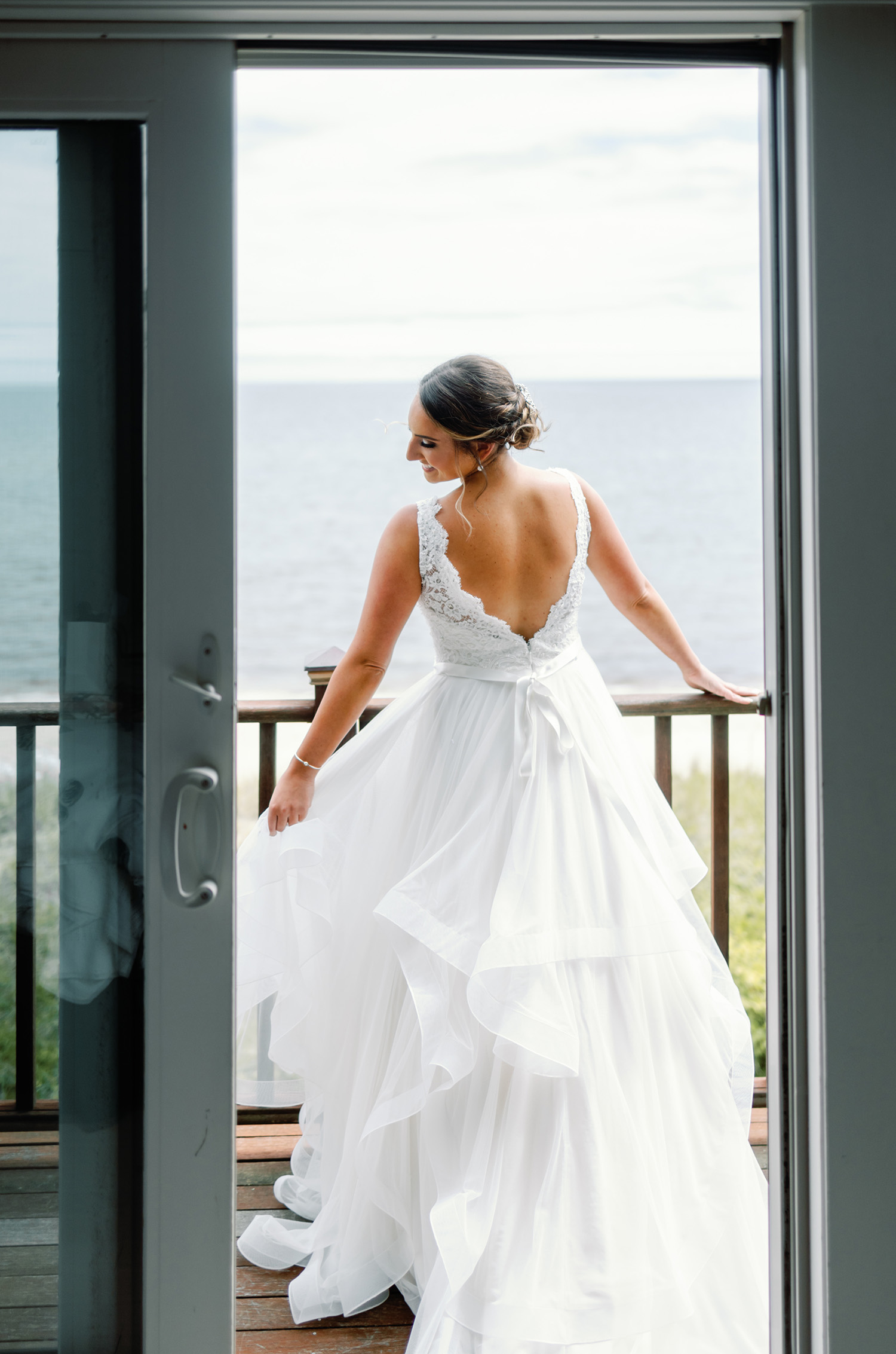  Bride poses on a balcony with an oceanview, in a open back lace gown with a multi-layered tulle skirt and ribbon at the waist. 