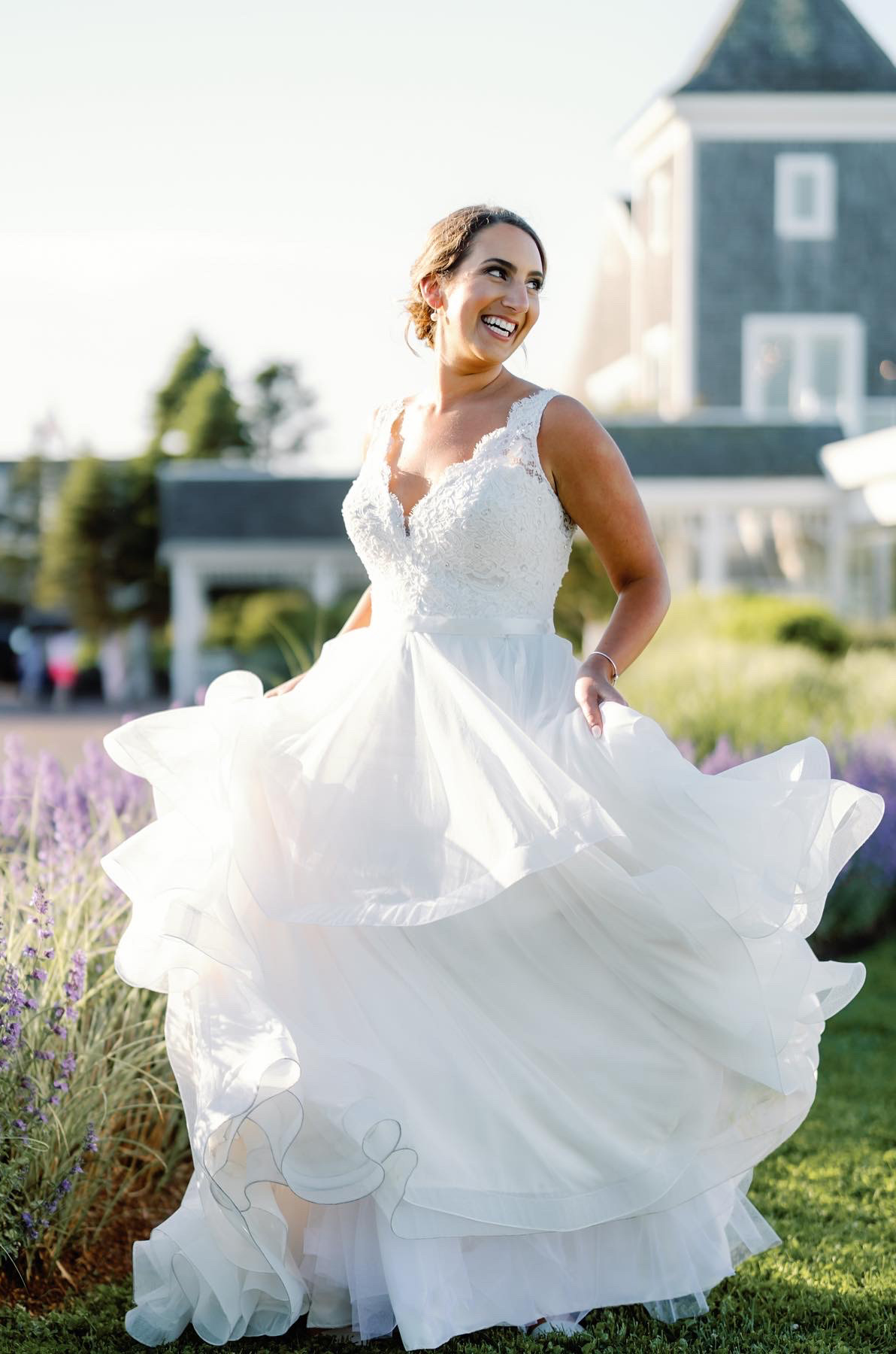 Bride walks through a wildflower field smiling in a V-neck lace gown with ribbon and a ruffled tulle skirt, near venue building. 
