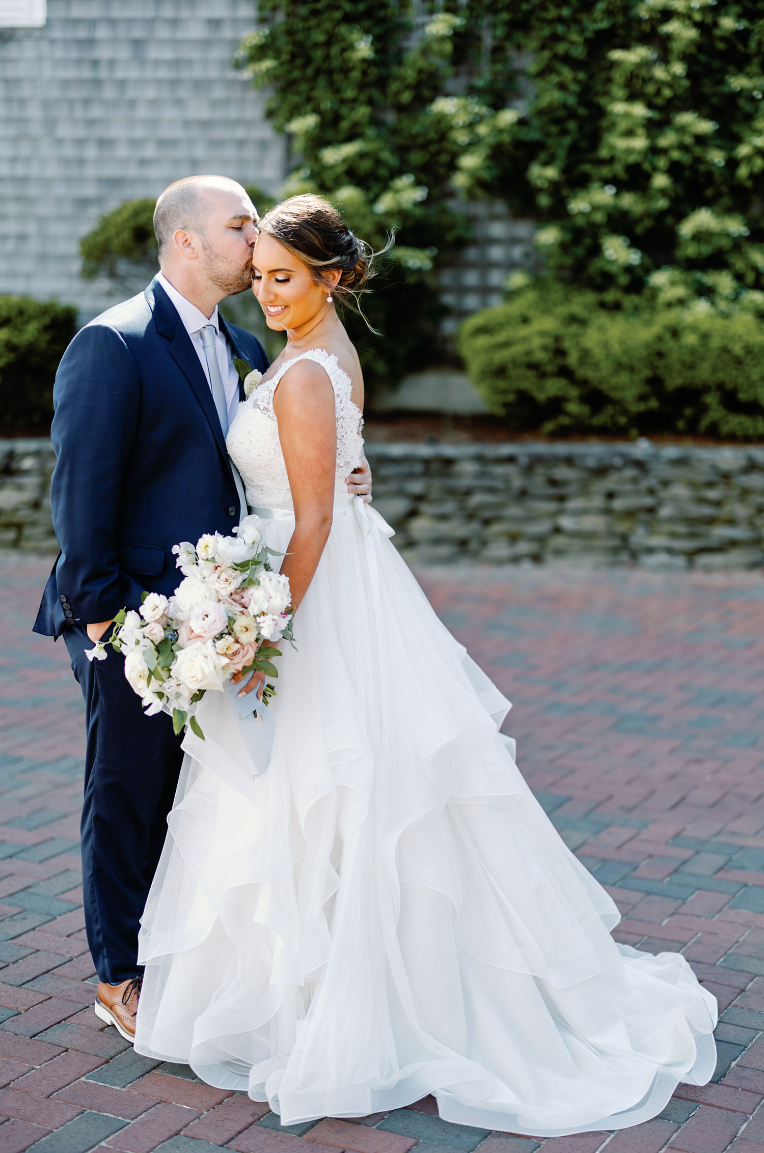 Groom embraces bride in a sleeveless lace and tulle gown with a ruffled skirt and ribbon, holding a bouquet in a courtyard.