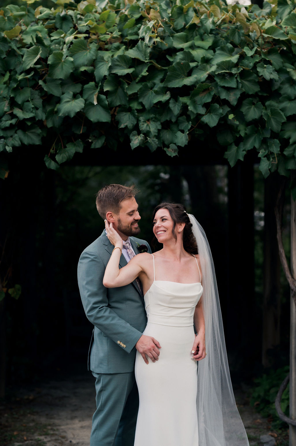 Close-up of groom embracing bride in a fit and flare crepe gown with a scoop neckline and a veil at a garden arbor with leaves.