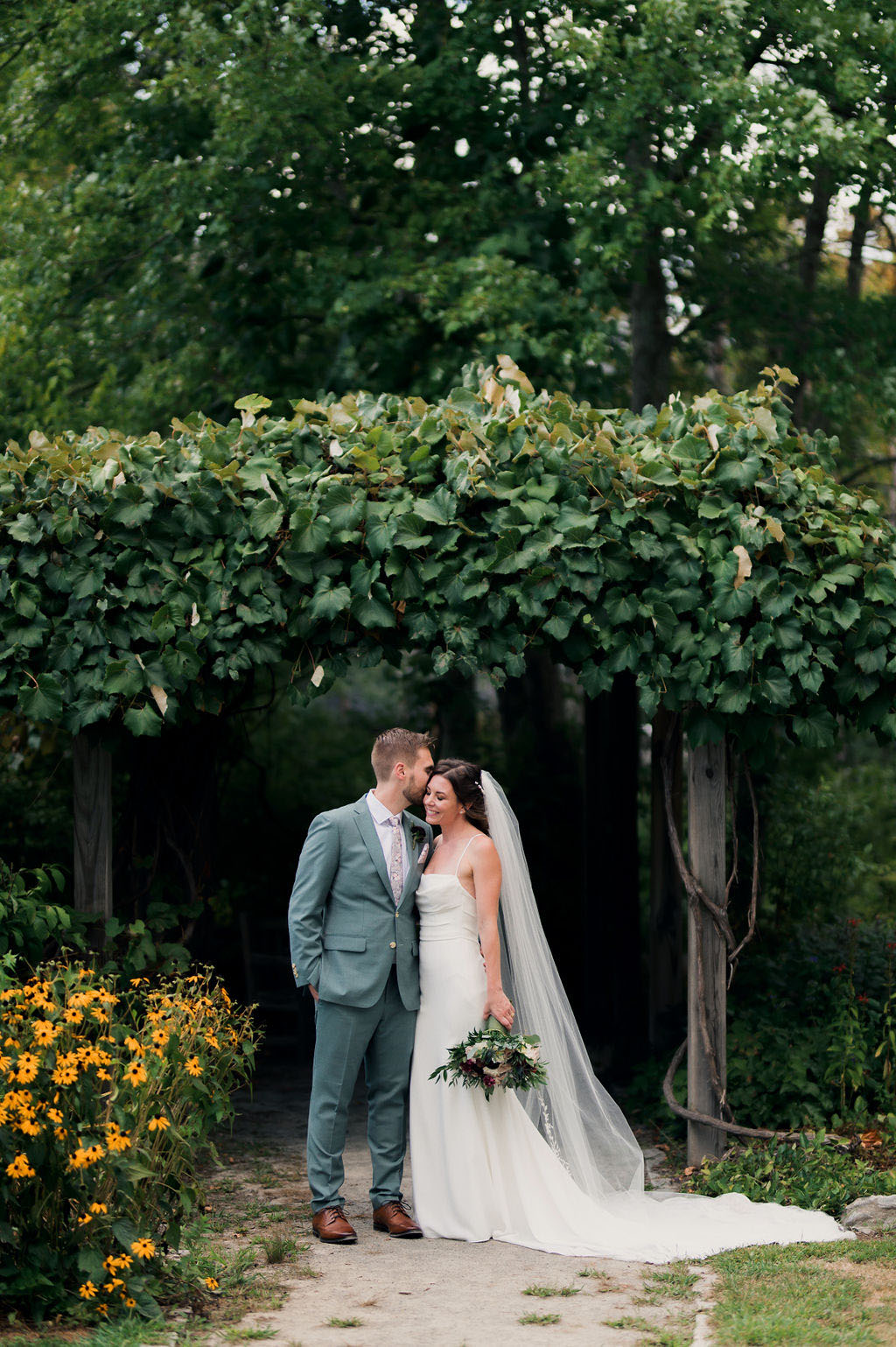 Bride in a crepe fit and flare wedding dress with a scoop neck, thin straps and a veil, poses with groom at a garden pergola.