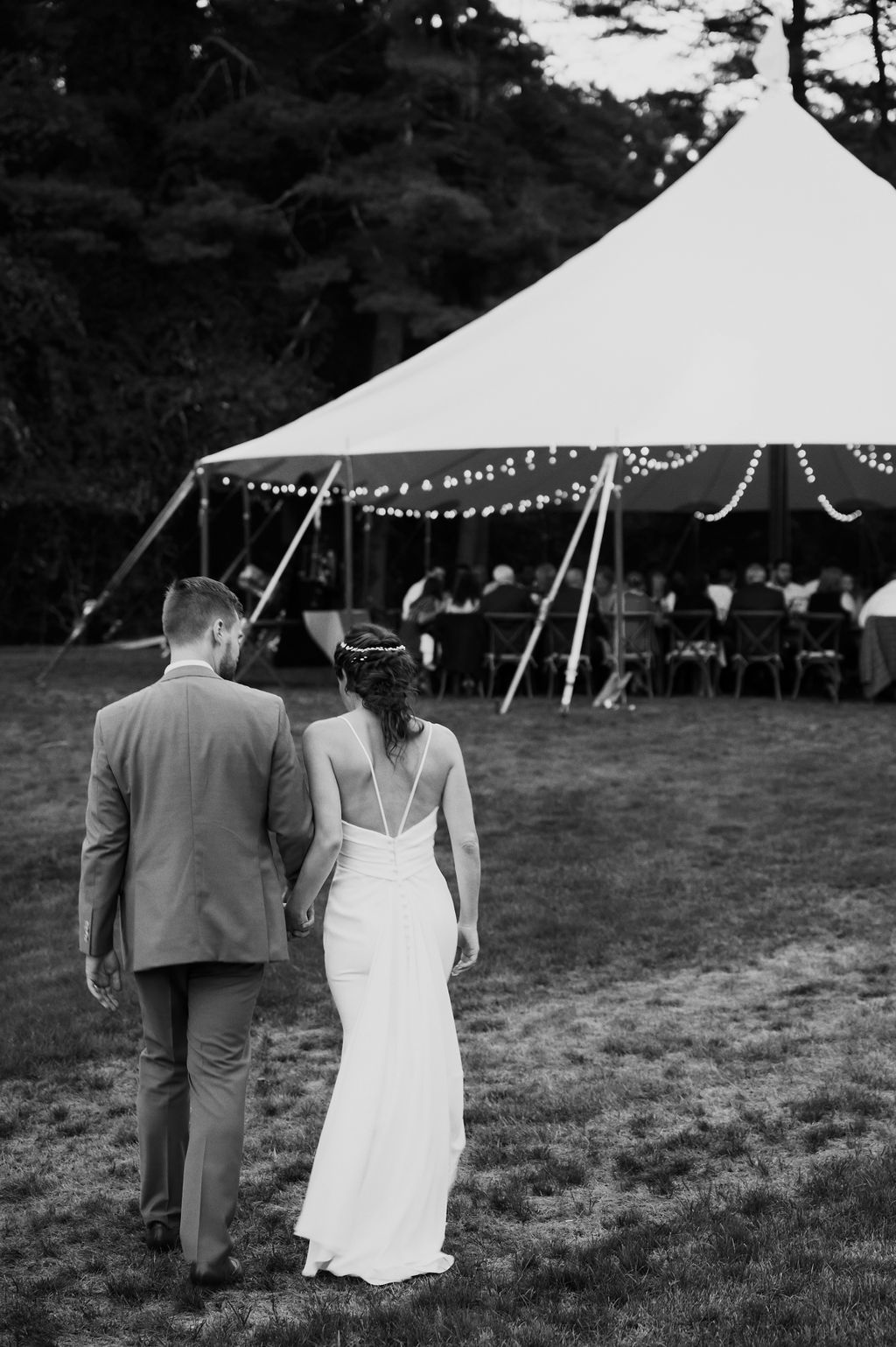 Back of bride in a spaghetti strap crepe fitted gown, walks alongside her groom, toward tented reception, in black and white.