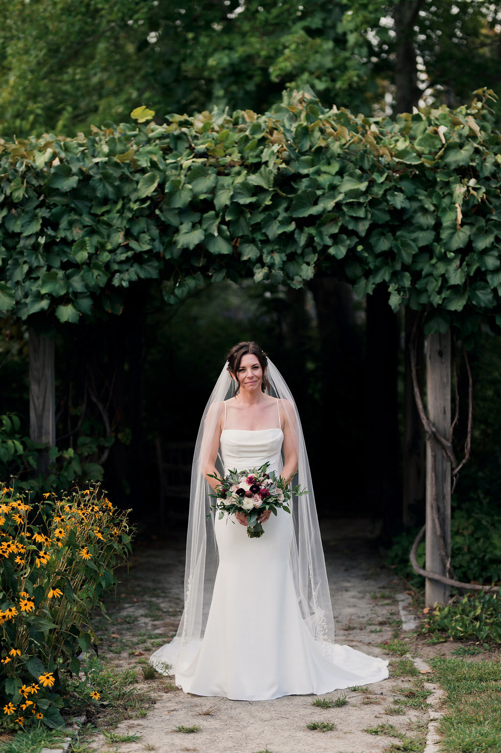Bride poses under a garden pergola in a scoop neck crepe gown with fit and flare skirt, veil and pleating, holding a bouquet.