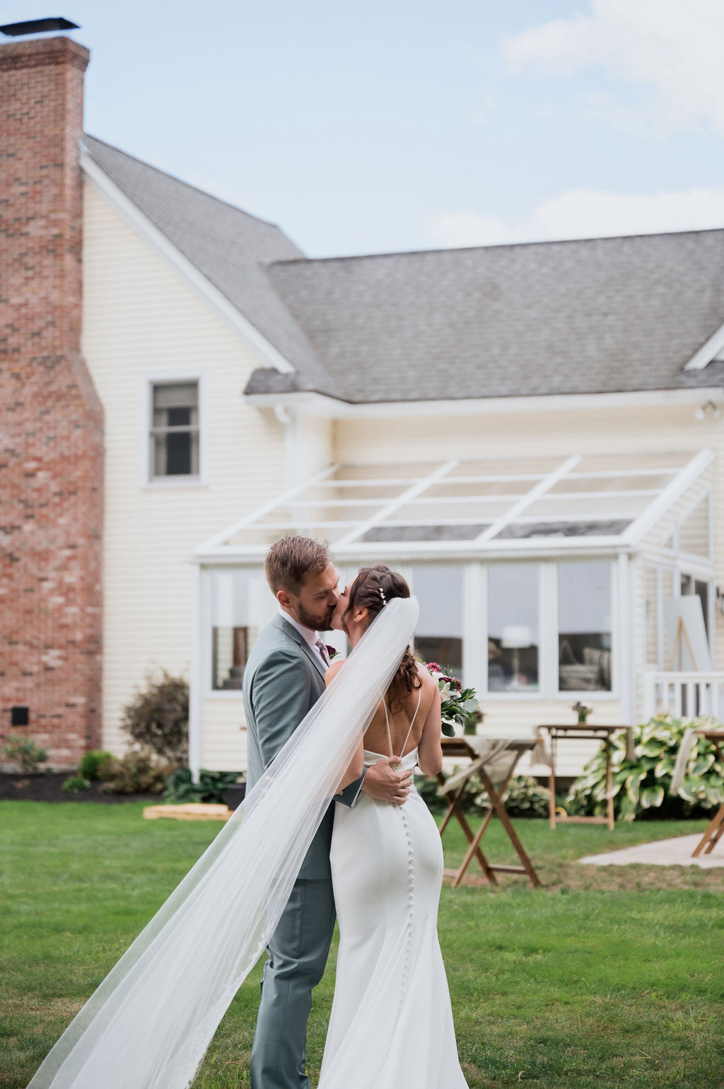 Groom embraces his bride in a low back crepe gown with a buttoned fit and flare skirt and a veil, as they kiss by a house.