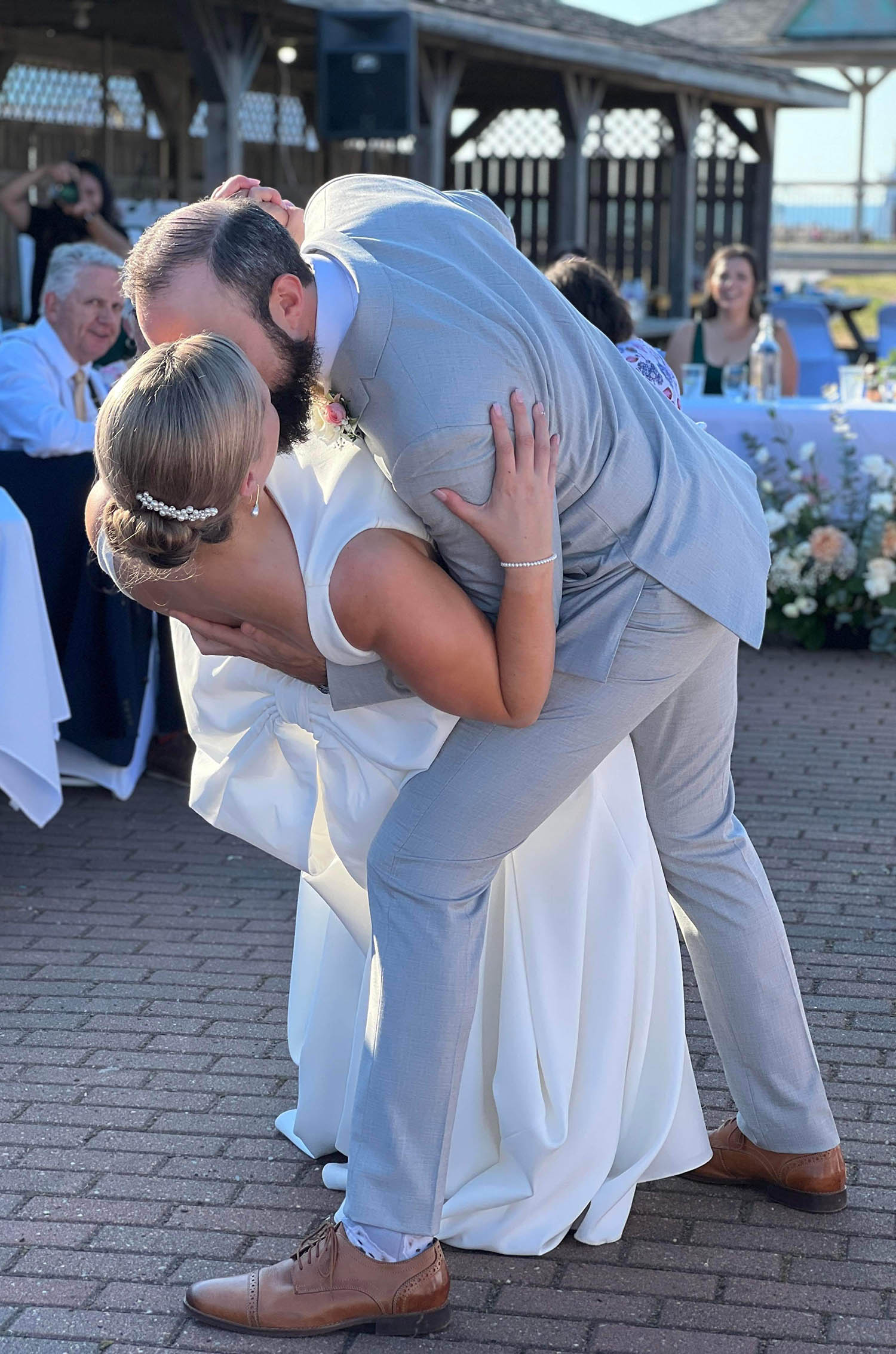 Groom dips his bride in a bateau neck crepe wedding dress with a bow at the back, at outdoor ceremony in front of guests.