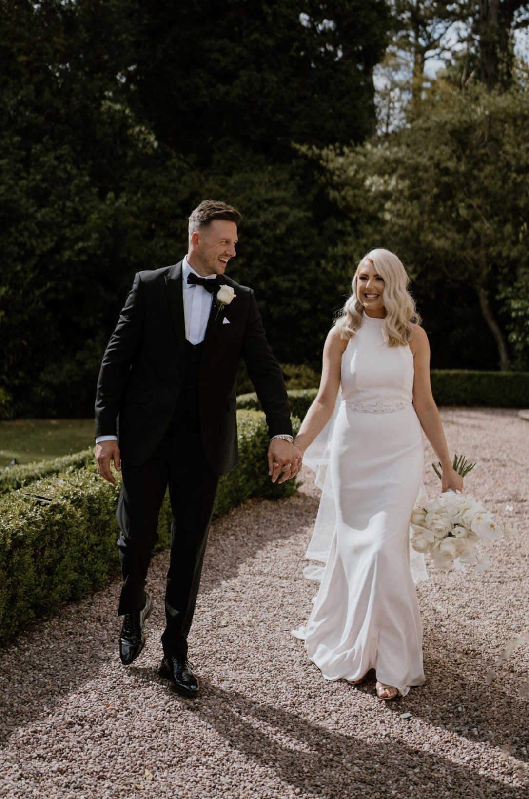 Bride walks in a high neck crepe fitted gown with belt detail, beside her groom in a tux, holding a white bouquet outdoors.