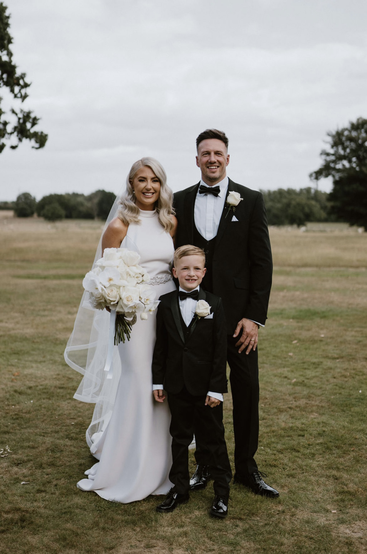 Bride in a high neck crepe gown with a beaded belt and veil, poses beside groom and pageboy on an estate lawn. 