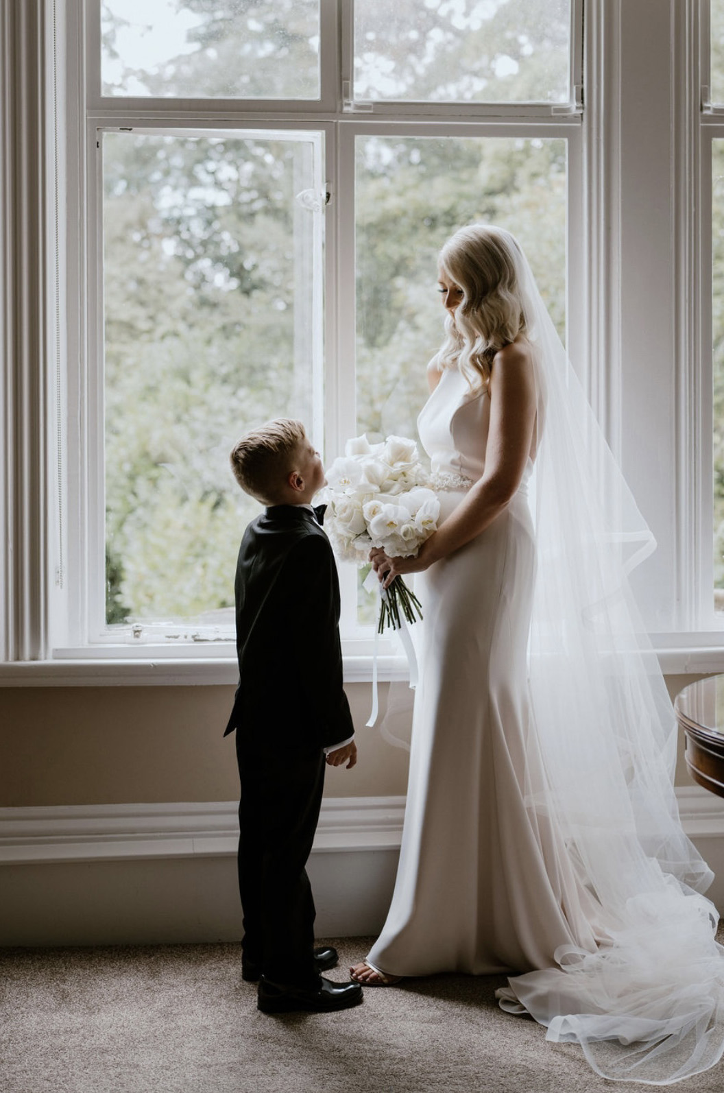 Bride in a halter neck crepe fitted gown is holding a white bouquet, and is posing at a bright open window. 