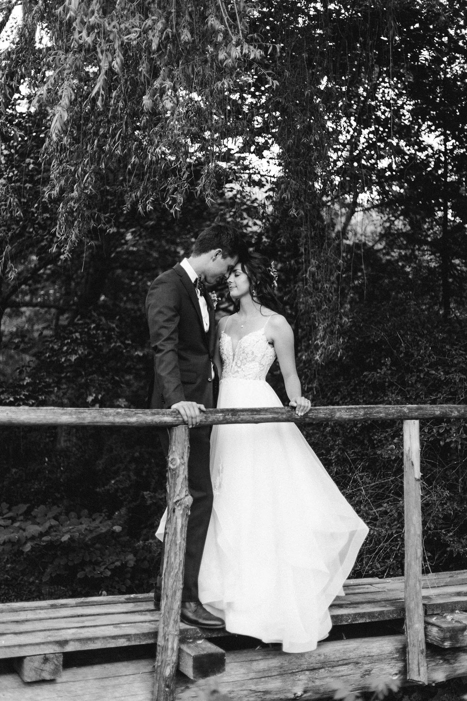 Bride in a spaghetti strap lace and organza gown, poses with groom under willow trees, on a wooden bridge in black and white.