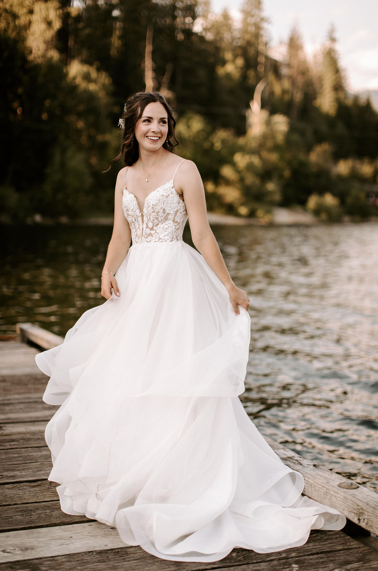 Bride twirls in a sleeveless lace bodice wedding dress with a flowing organza skirt, on a lakeside wooden dock in a forest. 