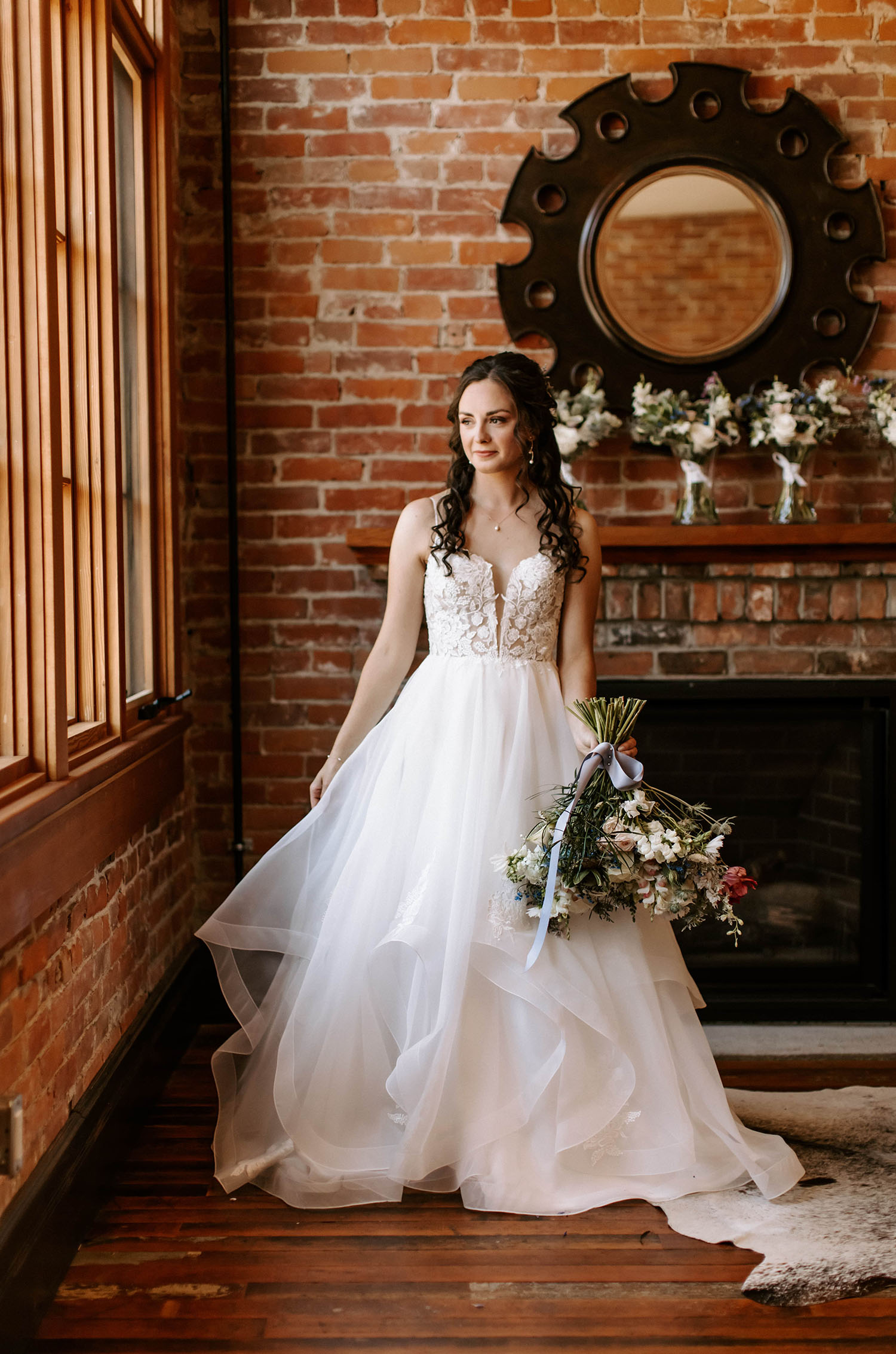 Bride poses by a brick fireplace in a lace gown with a plunging neck and tiered organza skirt, is looking out a window. 