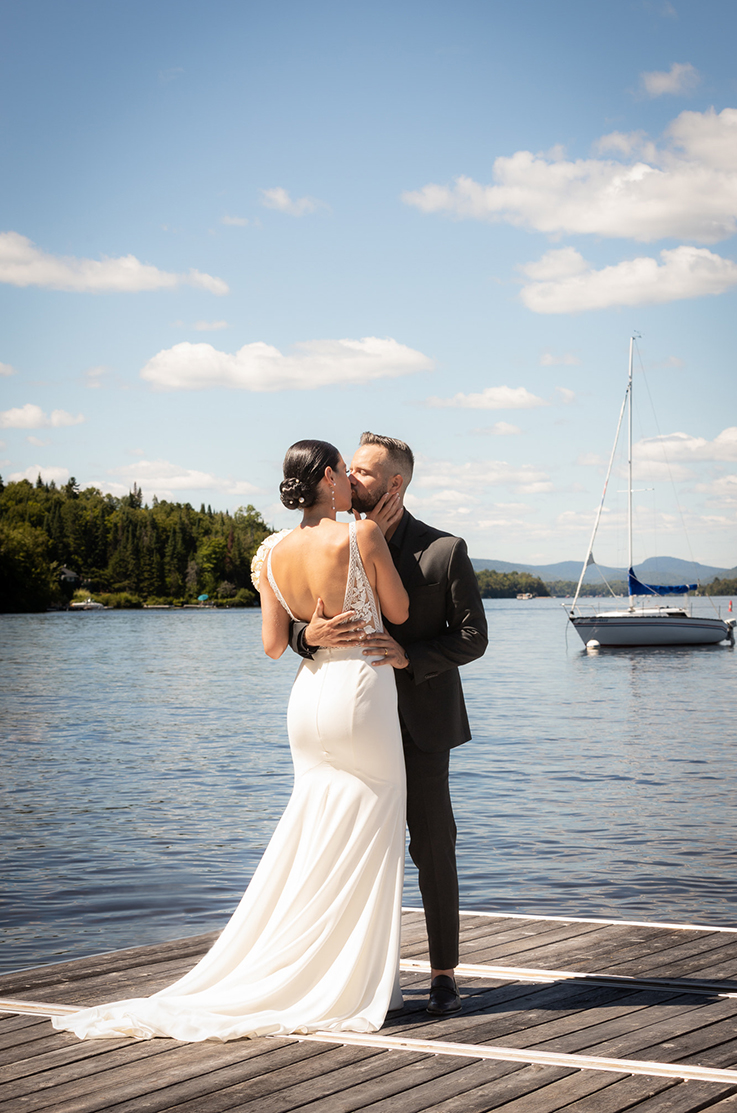 Groom kisses his bride in an open back lace gown with a crepe fit and flare skirt, on a lakeside dock near a sailboat.