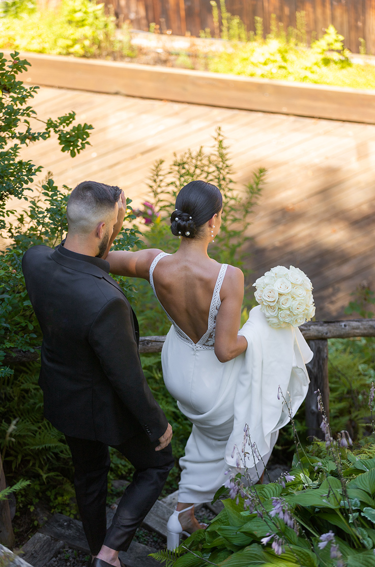 Groom walks down steps with his bride in a deep-V back lace gown with a crepe skirt, holding her train and a white bouquet. 