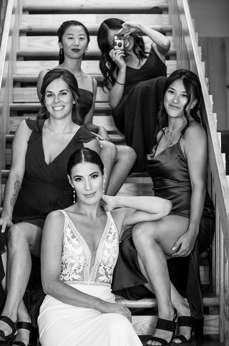 Bride in a plunging V-neck lace and crepe gown, sits on a staircase and poses with her bridesmaids, in black and white.