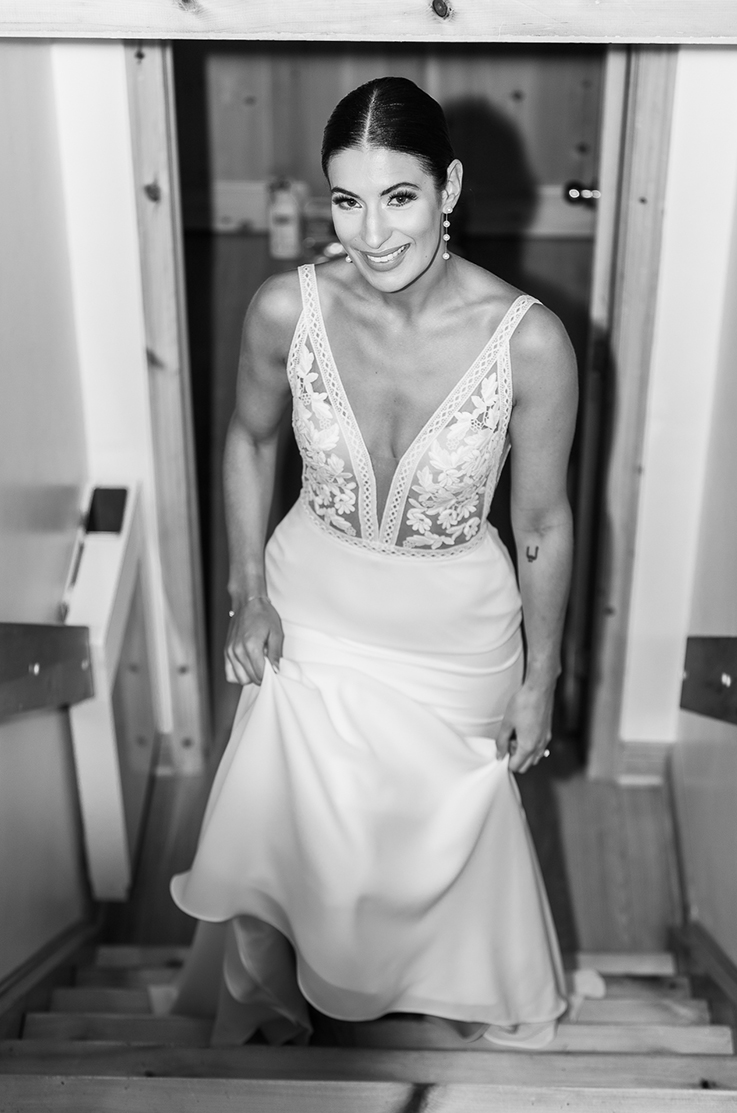 Bride walks up steps in a V-neck lace bodice gown with a fitted crepe skirt and picks up her train, in black and white.