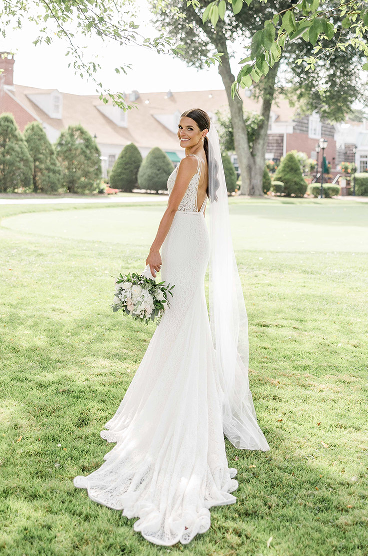 Bride looks over her shoulder in a sleeveless lace fitted gown with a deep-V back and a long veil, poses on a green lawn.