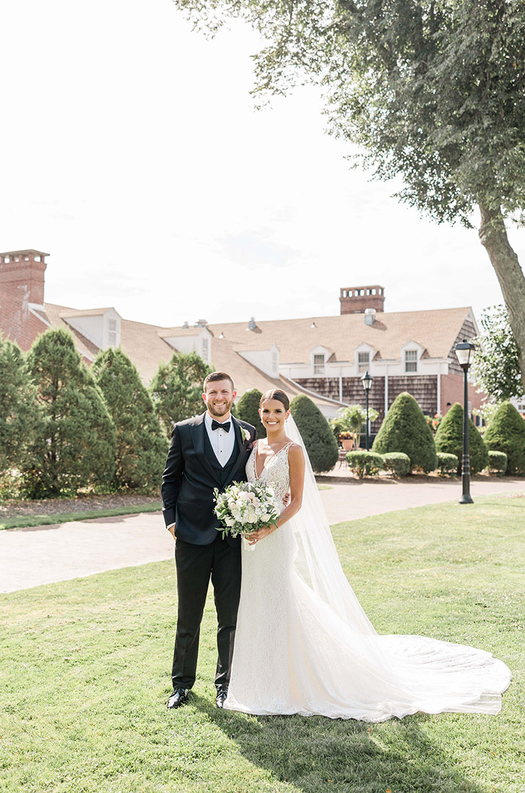 Bride in a fit and flare lace wedding dress with a V-neck and a long veil, poses next to groom in front of the venue. 