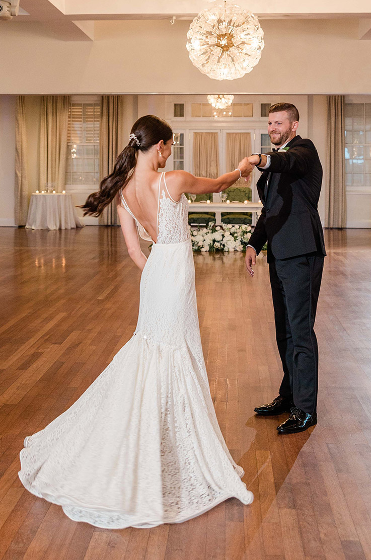 Groom dances with bride in a fit and flare lace gown with a low open back, in a chandelier-lit ballroom.