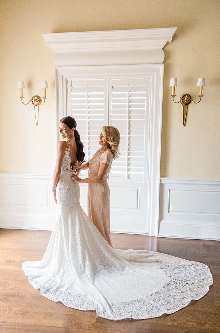 Bride's mother fastens bride into a sleeveless lace gown with a low back and fit and flare skirt, in a brightly lit room. 