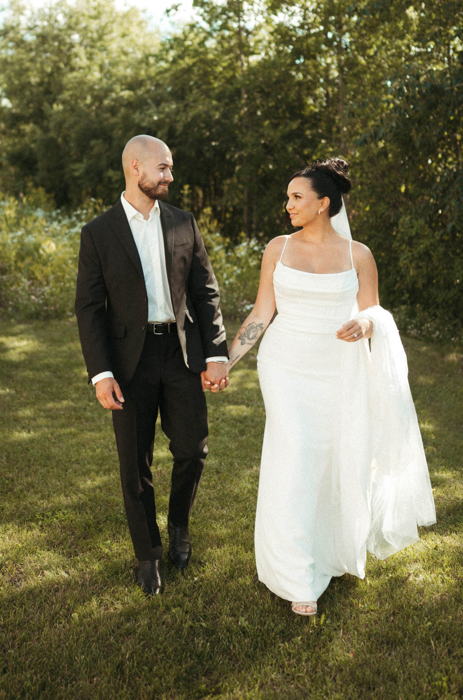 Bride in a scoop neck crepe gown with fit and flare skirt and a veil, holds the train and walks hand in hand with her groom.