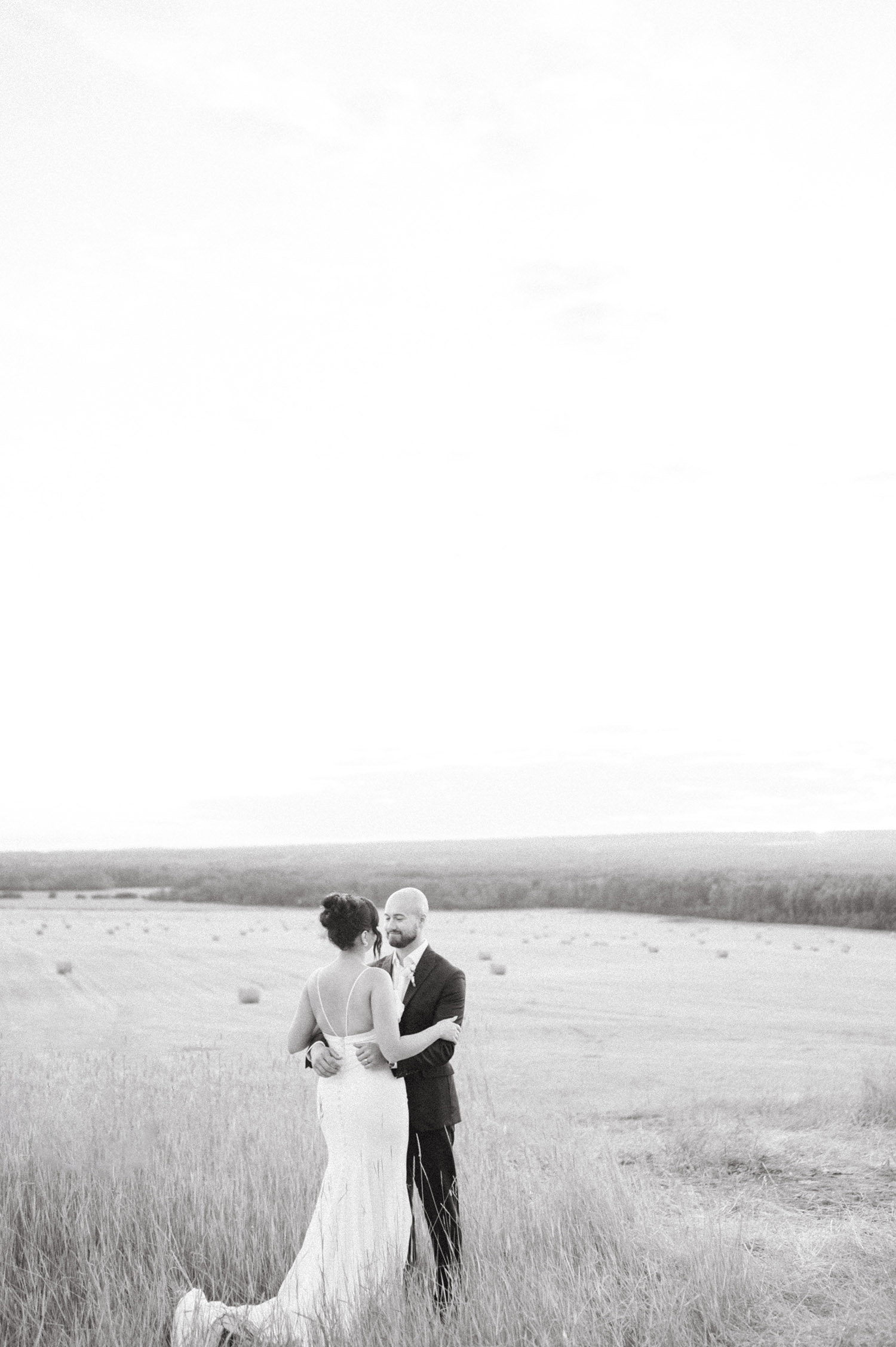  Groom embraces his bride in a crepe fit and flare wedding dress with a strappy low back, standing in an open field. 