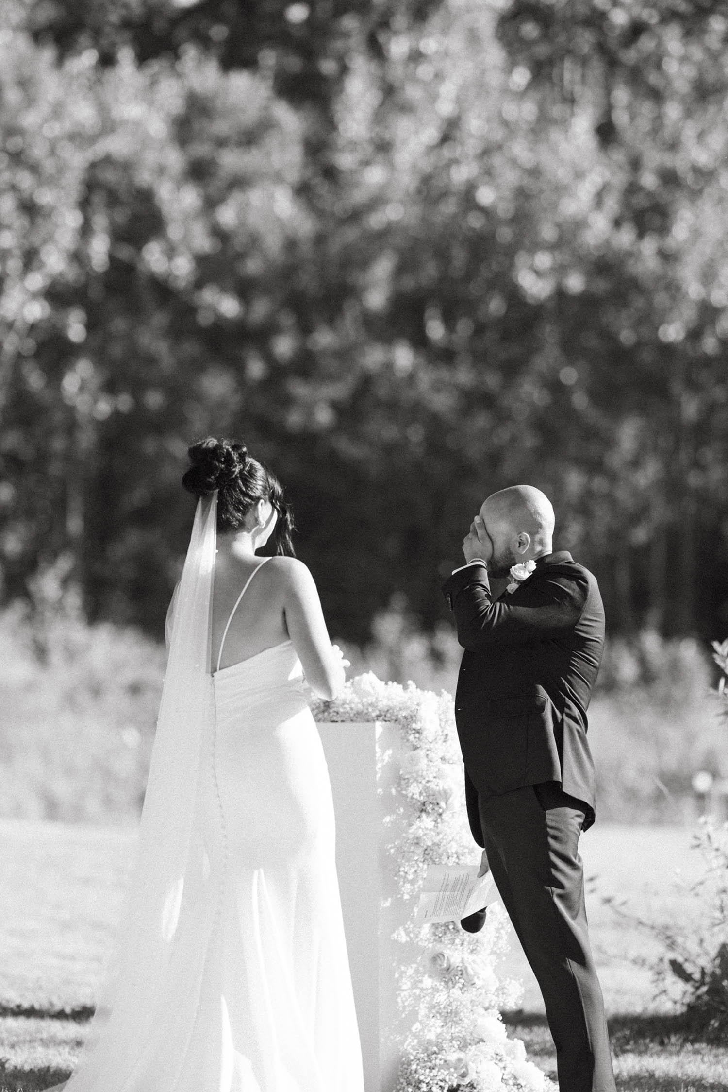 Bride in a crepe low back gown with buttons, a fit and flare skirt, thin straps and a veil, stands near groom under trees. 