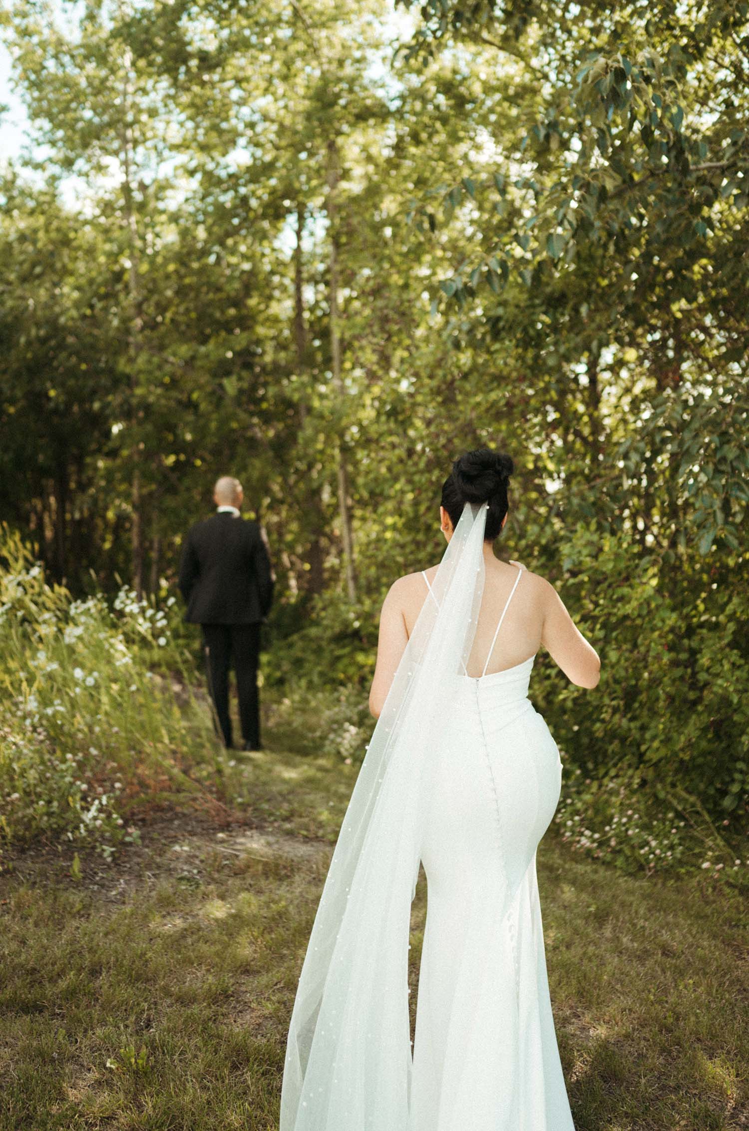 Bride in a fitted crepe gown with a buttoned low back, spaghetti straps and a pearl veil, walks towards the groom in grass. 