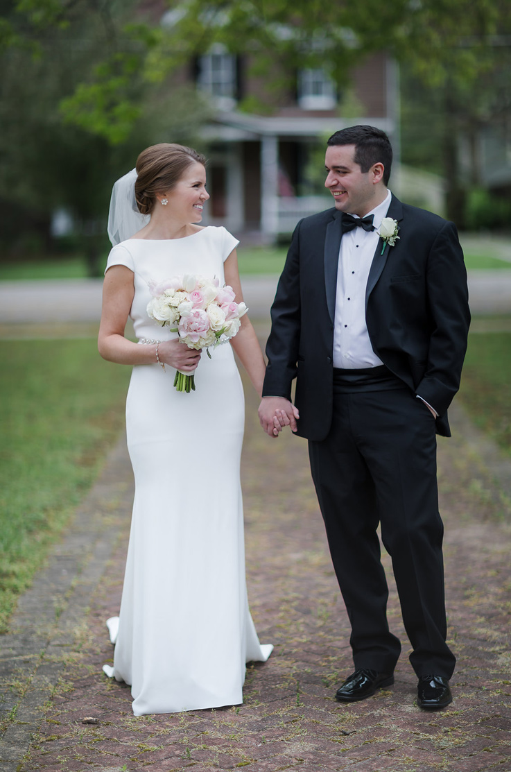 A bride in a cap sleeve wedding dress and veil, holding a bouquet in one hand, holds the hand of a groom in a suit while they smile and look at each other outdoors on a gravel path.