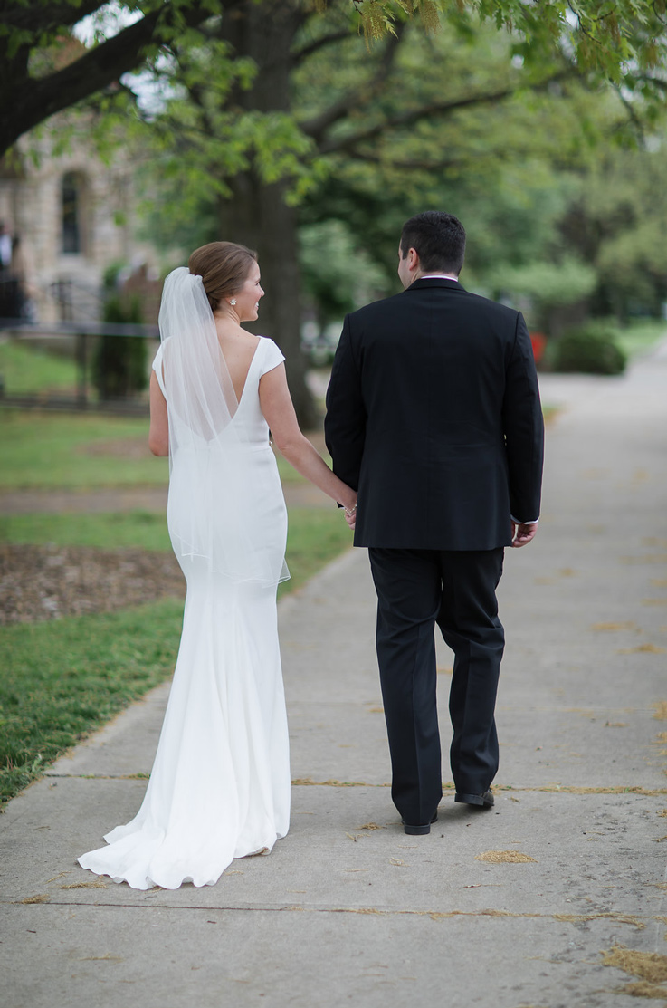 A bride in a cap sleeve wedding dress and veil, and a groom in a suit, walk down a path outdoors with a stone building and trees in the background.