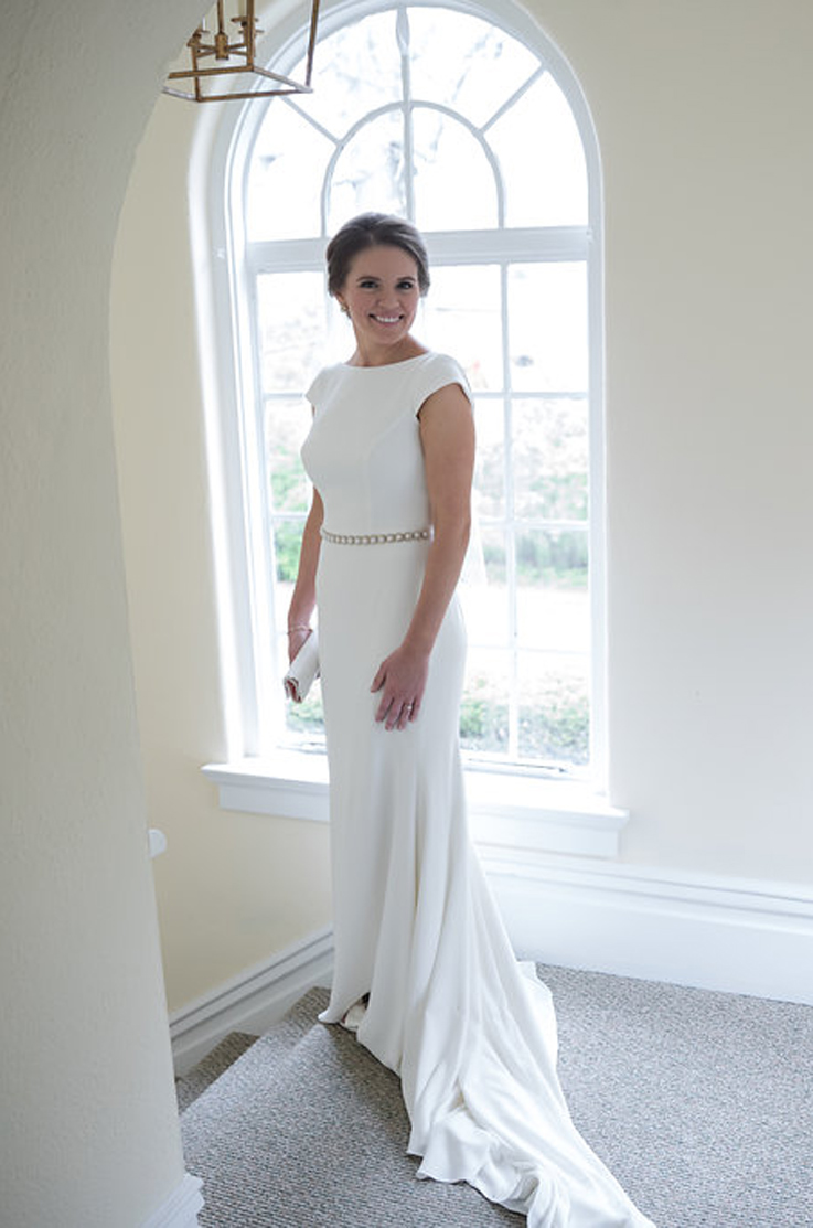 A bride in a cap sleeve wedding dress smiles and poses at the top of a staircase indoors, with an arched window behind her.