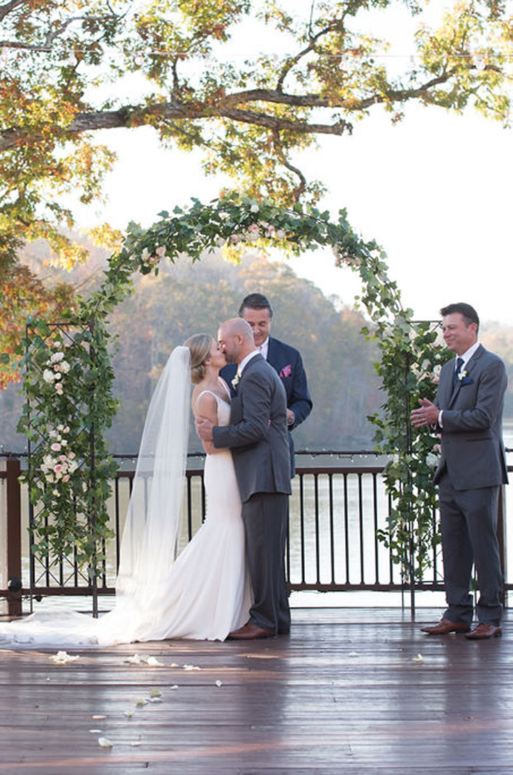 A bride in a fitted wedding dress and a groom in a suit kiss on a wooden outdoor patio, in front of a man in a suit, a floral wedding arch, and a lake.