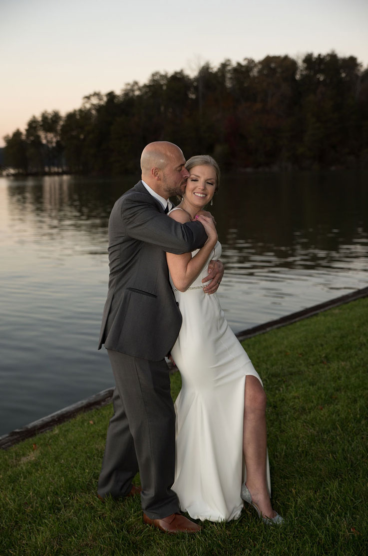 A groom in a suit kisses the cheek of a bride in a skirt slit wedding dress as she smiles, while standing on grass outdoors, in front of a lake with trees in the background.