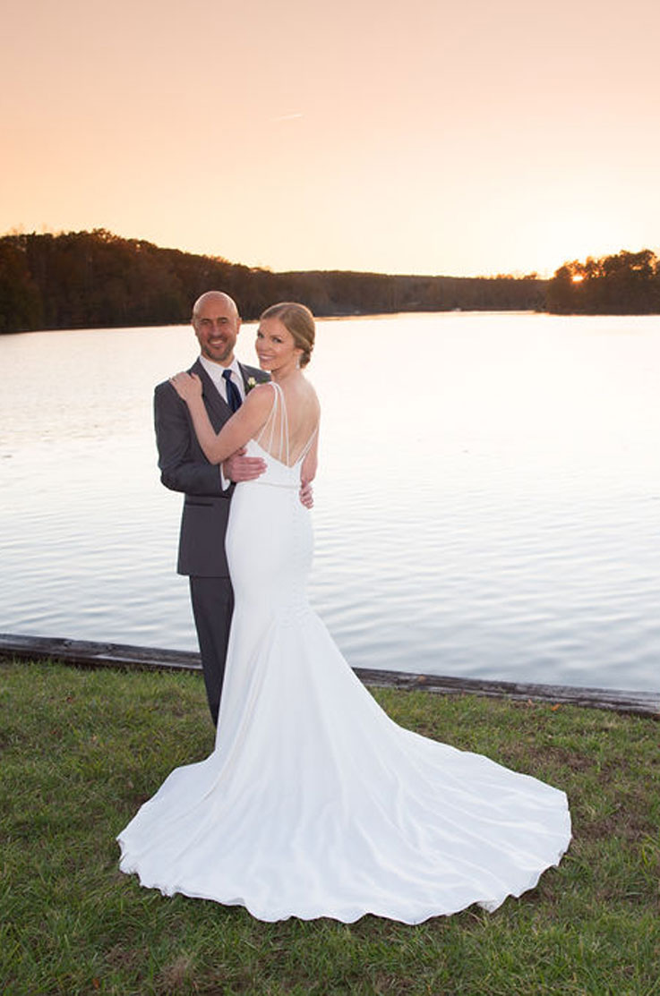 A bride in a fitted wedding dress and a groom in a suit hold each other and smile while standing on grass outdoors, in front of a lake.