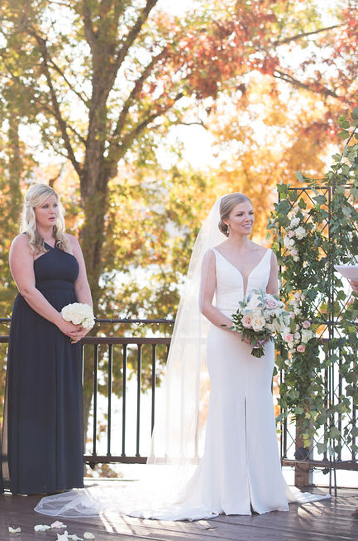 A bride in a plunging neckline wedding dress holds a veil while standing on a wooden outdoor patio, with a bridesmaid standing off to her side.