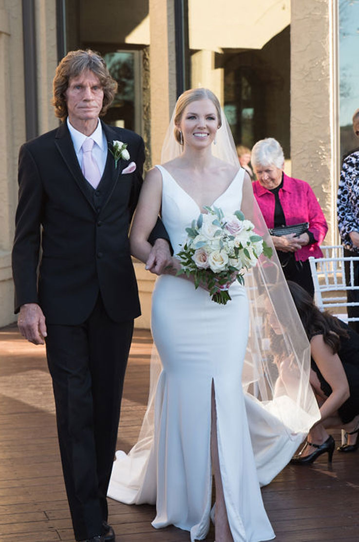 A bride in a skirt slit wedding dress and veil, holding a bouquet, links arms with a man in a suit as she smiles and walks forward, with wedding guests in the background.