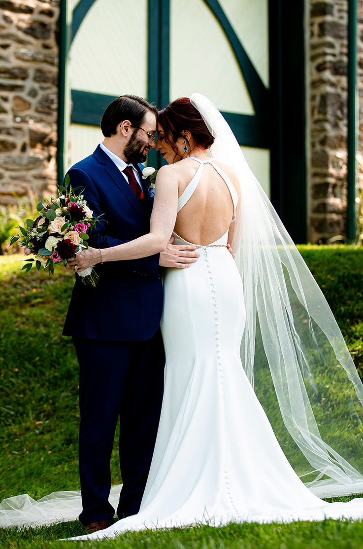 A bride in an open back wedding dress and a veil, holding a bouquet, stands close to a groom in a suit while outdoors on a grass lawn in front of a stone building.