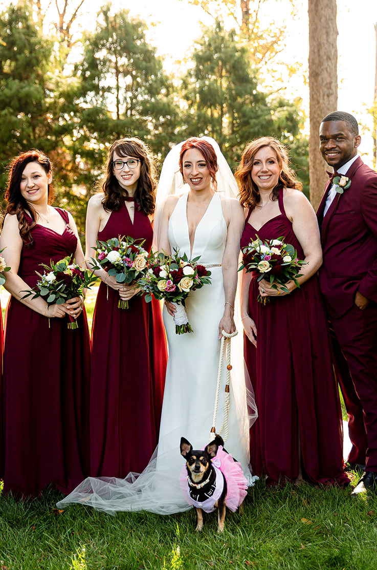 A bride in a plunging neckline wedding dress smiles and holds the leash of a small sweet dog, while standing between three bridesmaid and a man in a suit, while holding bouquets and standing outdoors.