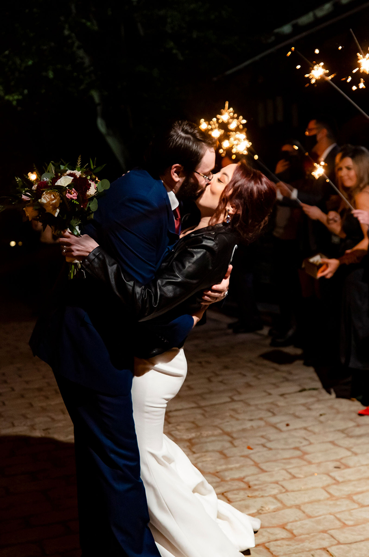 A groom in a suit dips and kisses a bride in a fitted wedding dress and leather jacket, while standing outdoors in the dark, with wedding guests holding sparklers stand behind them.